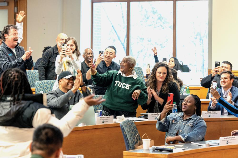 A group of Tuck Entrepreneurial Impact Program participants stand in a Tuck classroom in Hanover, NH, clapping. They are applauding for a fellow participant during the Building a Successful Business program graduation ceremony.